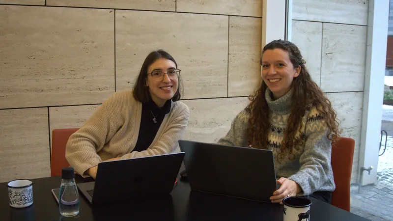 Elena (a sinistra) e Giulia (a destra) sedute a un tavolo con laptop e tazze di caffè