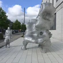 Voluminosa scultura argentata e figura umana metallica lungo un marciapiede