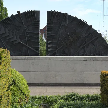 Scultura astratta in bronzo a forma di ventaglio su muro di pietra
