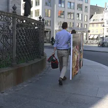Uomo cammina lungo un marciapiede portando un grande dipinto colorato