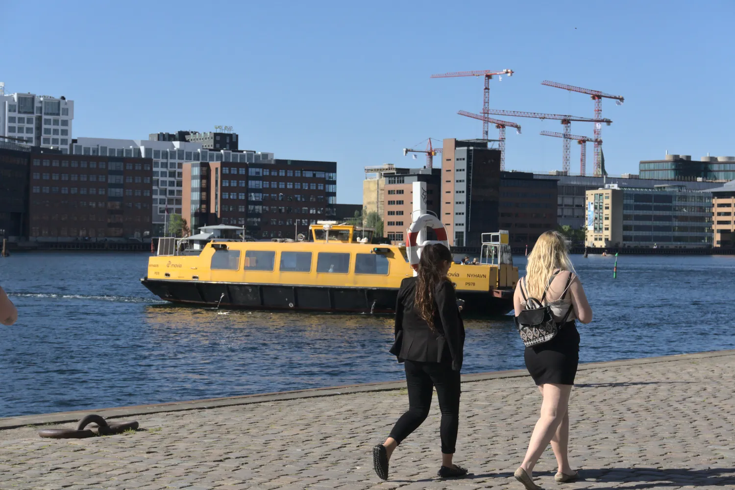 Due donne camminano sulla banchina mentre un battello giallo attraversa il porto