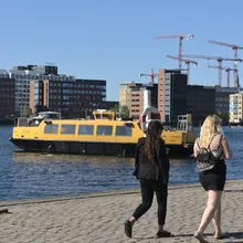 Due donne camminano sulla banchina mentre un battello giallo attraversa il porto