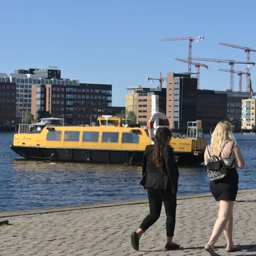 Due donne camminano sulla banchina mentre un battello giallo attraversa il porto