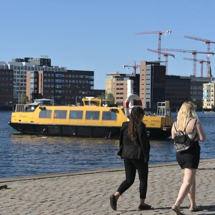 Due donne camminano sulla banchina mentre un battello giallo attraversa il porto