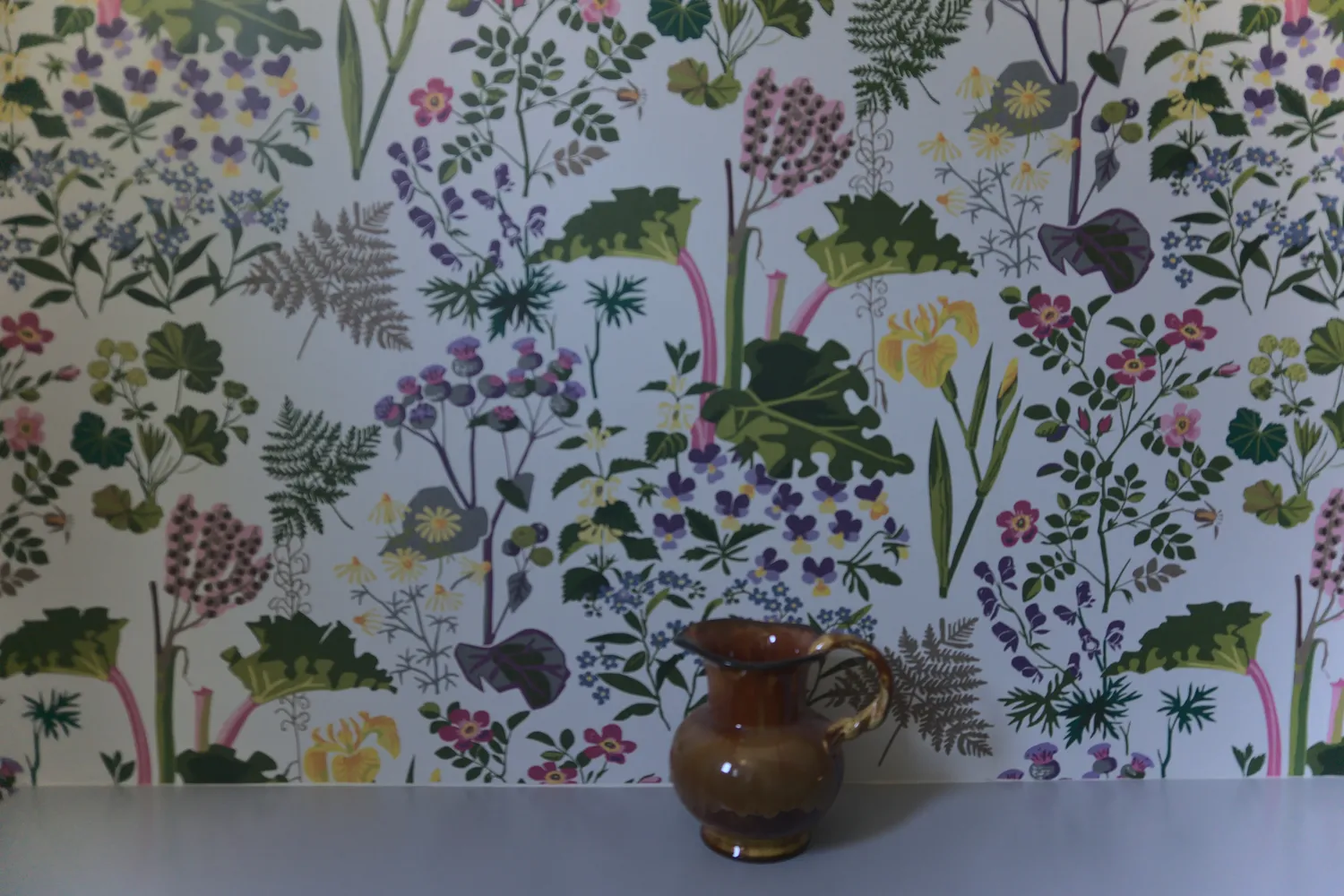Brocca di ceramica marrone davanti a una parete con carta da parati floreale
