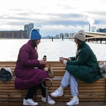 Due donne sedute su una panca di legno bevono caffè davanti al porto
