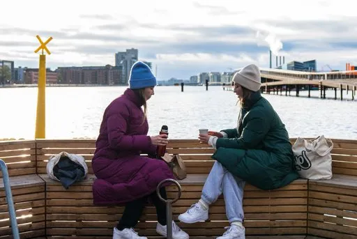 Due donne sedute su una panca di legno bevono caffè davanti al porto