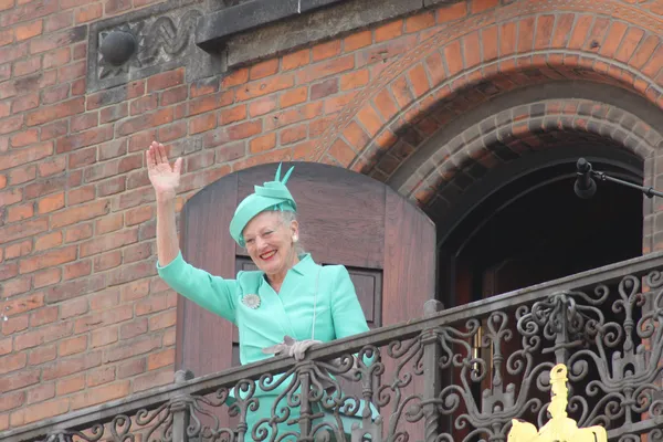 Donna anziana in abito verde saluta dal balcone di un edificio in mattoni