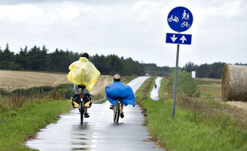 Due ciclisti con mantelle impermeabili su una pista ciclabile tra campi danesi