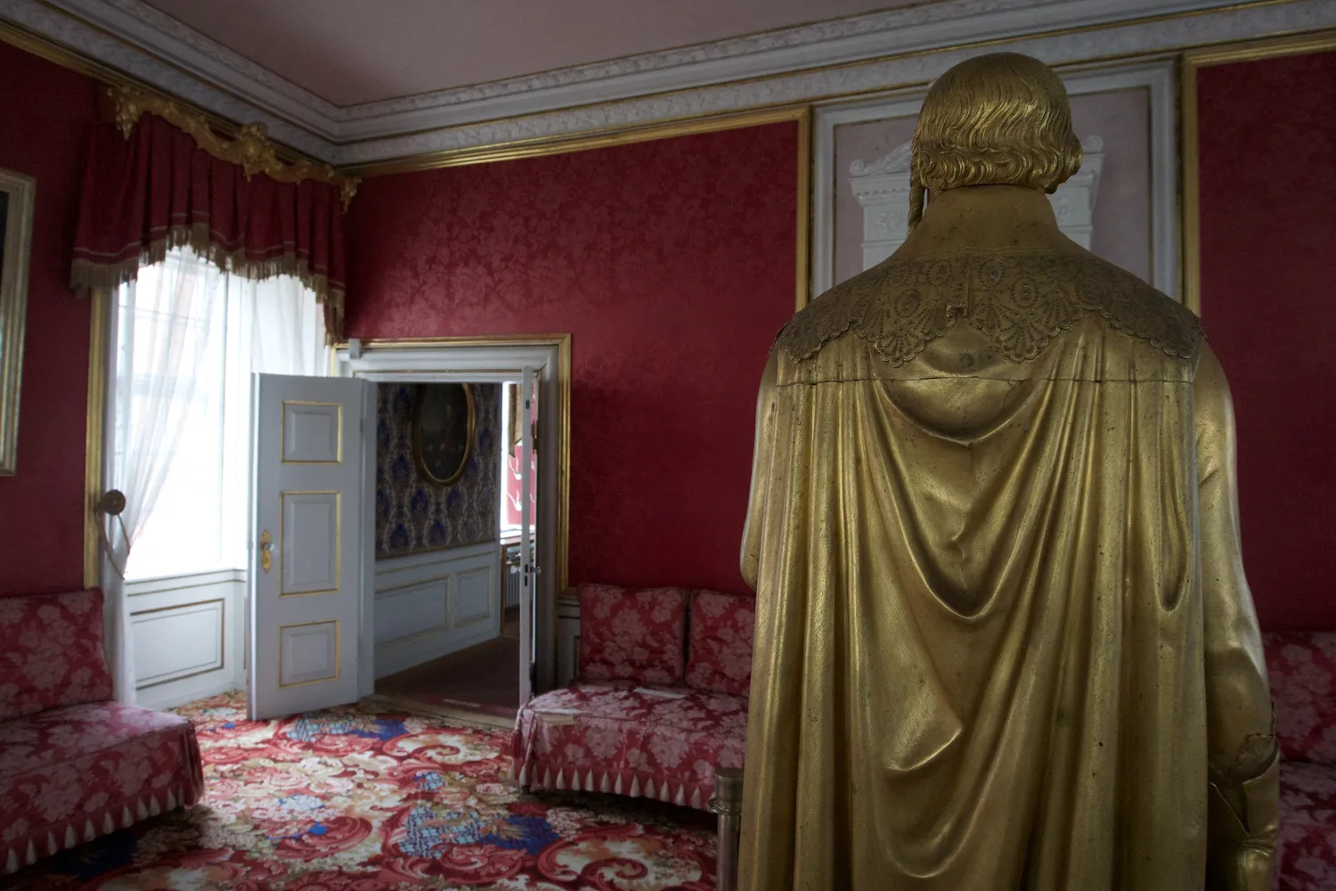 Statua dorata vista di spalle in una sala con pareti rosse damascate